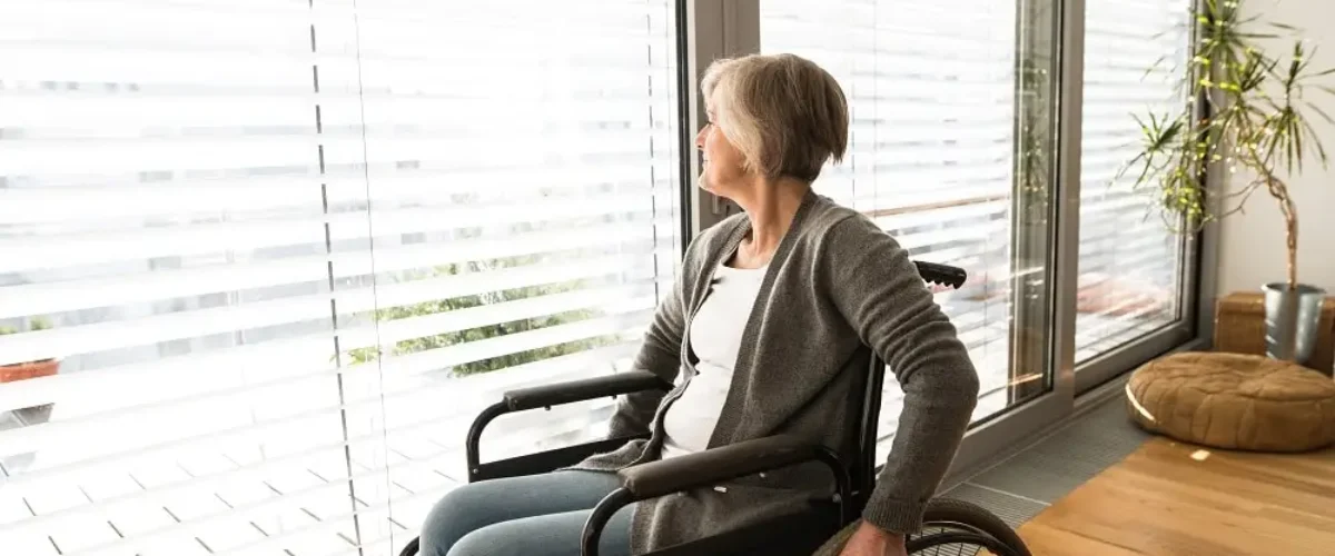 Disabled senior woman in wheelchair at home in her living room, looking out the window.